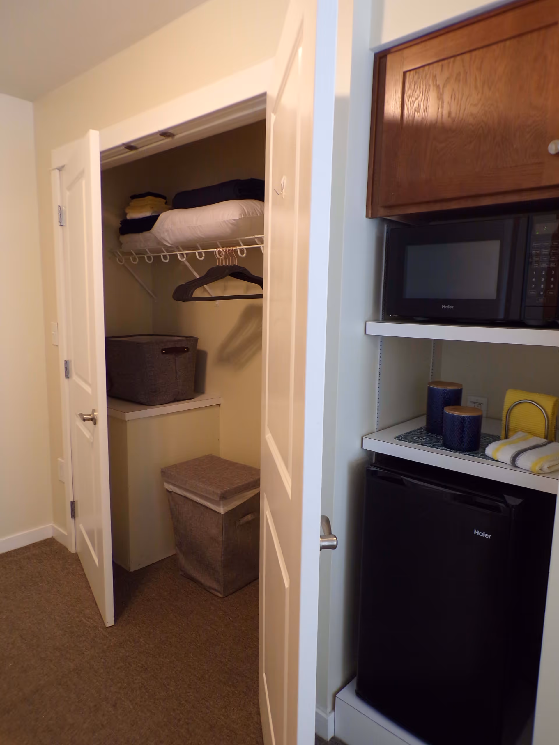 A small closet with white double doors open, showing folded linens and towels on a wire shelf, a gray storage bin on a white cabinet, and a gray hamper on the floor. To the right of the closet is a kitchenette area with a black mini fridge, a microwave, and two blue cups on a tray with a yellow and white striped cloth.