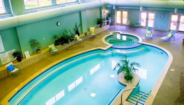 Indoor swimming pool area with a connected hot tub, surrounded by lounge chairs and potted plants. The room has large windows and glass doors letting in natural light, with a greenish wall color and a clock on the wall.