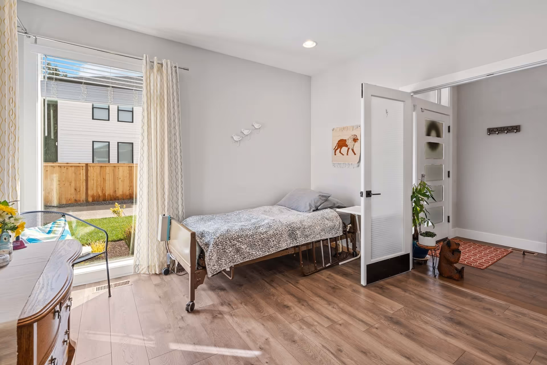 Sunlit bedroom with a single adjustable bed by a large window, wooden floors, and an open door to a hallway.