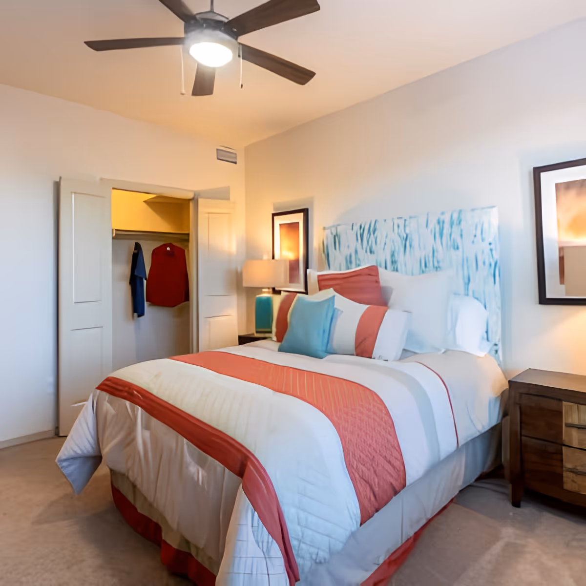 A neatly made bed with a colorful striped bedspread and multiple pillows in a bedroom. There is a ceiling fan with a light above the bed, a nightstand with a lamp, framed artwork on the walls, and an open closet with clothes hanging inside.