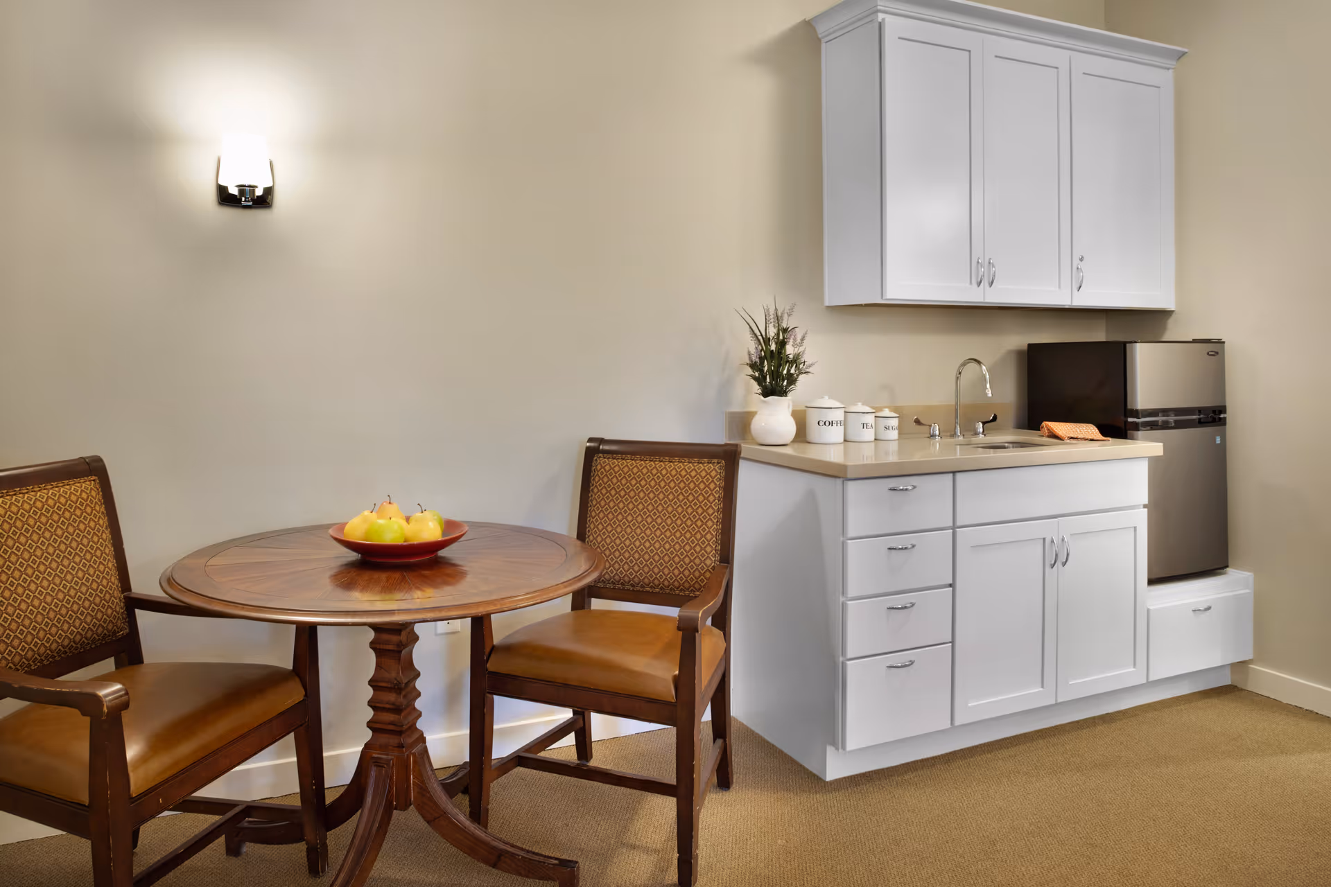 Small kitchenette and dining area with a round wooden table and two chairs beside white cabinets, a sink, and a mini fridge.