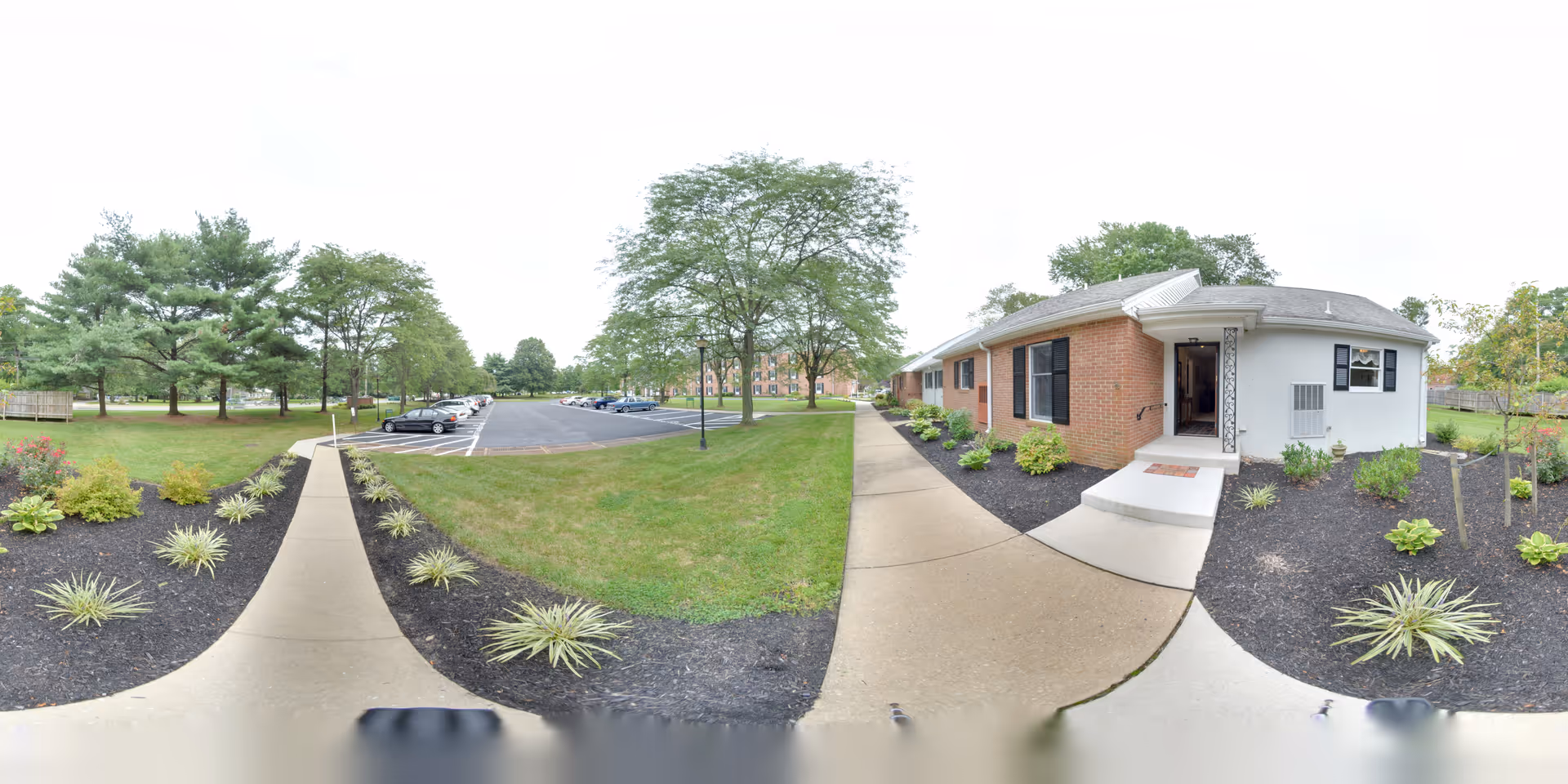Outdoor view of a senior living facility showing a brick building with a white entrance door and steps, surrounded by landscaped garden beds with small plants and mulch. A concrete walkway leads to the entrance, and there is a parking lot with several cars and trees in the background under an overcast sky.