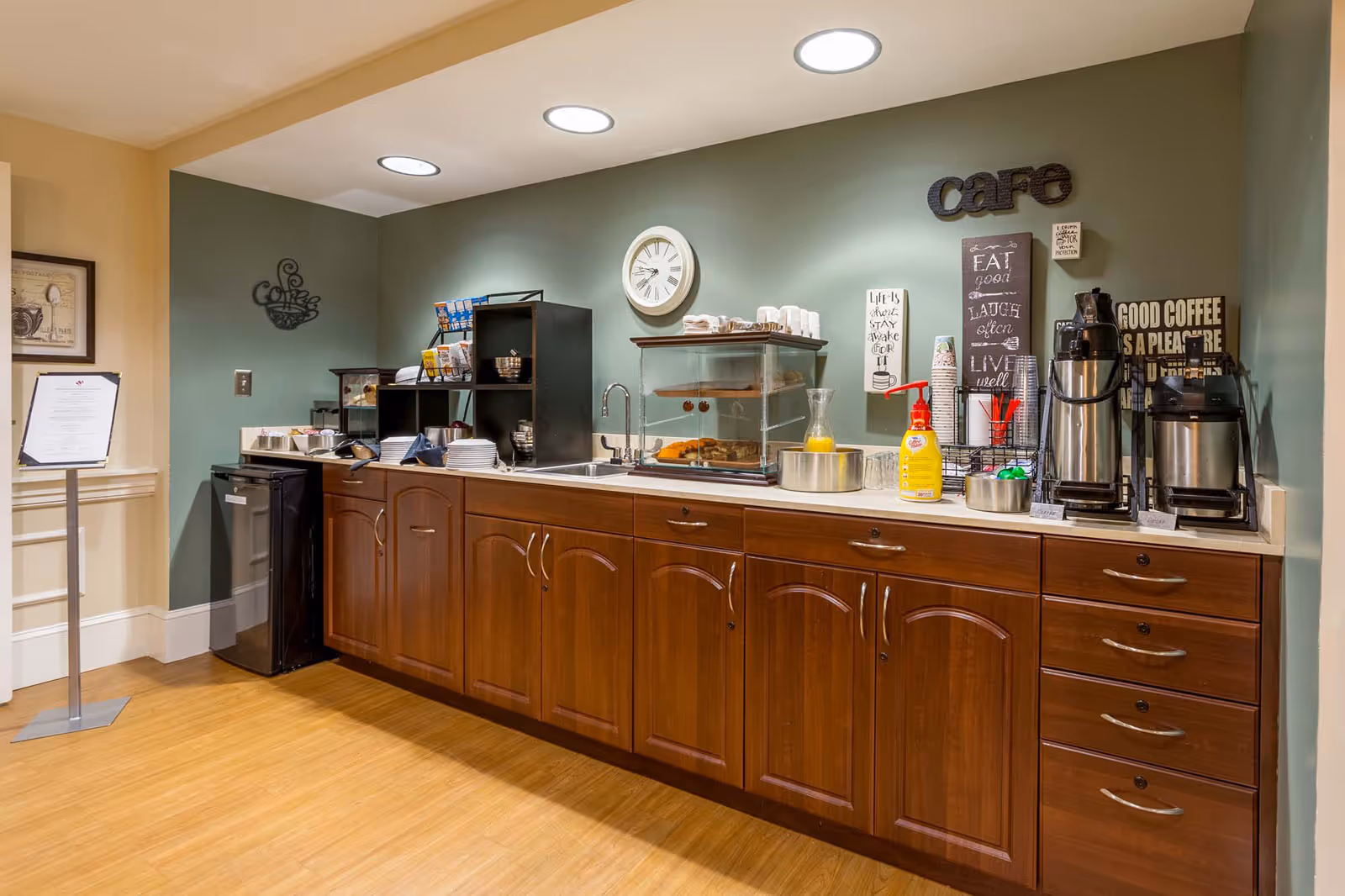 A coffee and snack service counter with wooden cabinets, coffee urns, a pastry display case, and wall decor reading "cafe".