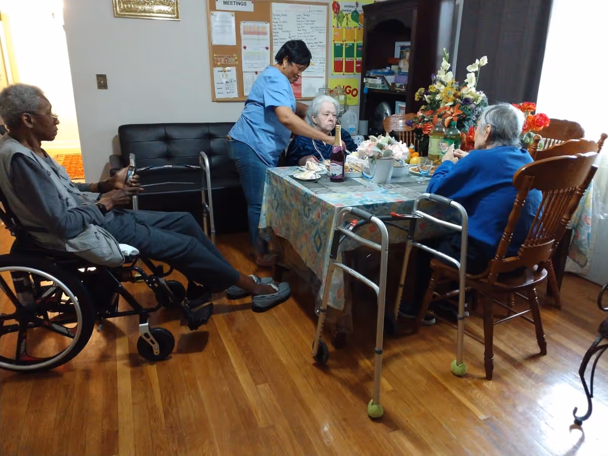 A caregiver in a blue uniform assists an elderly woman seated at a dining table covered with a patterned tablecloth. Two other elderly individuals are present, one in a wheelchair and the other seated on a wooden chair with a walker nearby. The room has wooden flooring, a black couch, a bulletin board with notes, and a cabinet with flowers and various items on top.