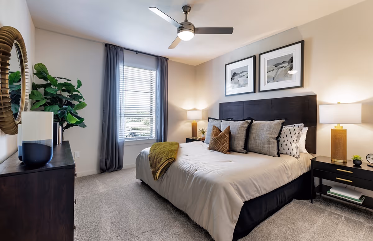 Well-lit modern bedroom featuring a large bed with multiple pillows, nightstands and lamps, a dresser with decor, a window with curtains, and a ceiling fan.