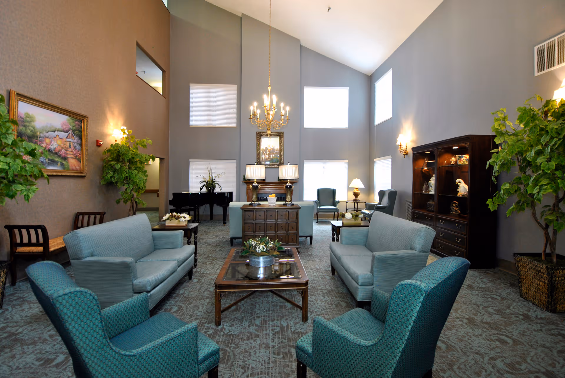 A spacious and well-lit living room with high ceilings, featuring two blue sofas, two blue armchairs, a glass coffee table with a plant centerpiece, a wooden cabinet with decorative items, and several lamps. There are large windows allowing natural light, a chandelier hanging from the ceiling, and a piano in the background. The room is decorated with plants and a framed painting on the wall.