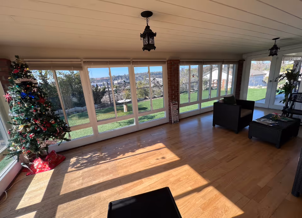 Sunlit enclosed living room/sunroom with large windows overlooking a yard, a decorated Christmas tree on the left and wicker seating on the right.