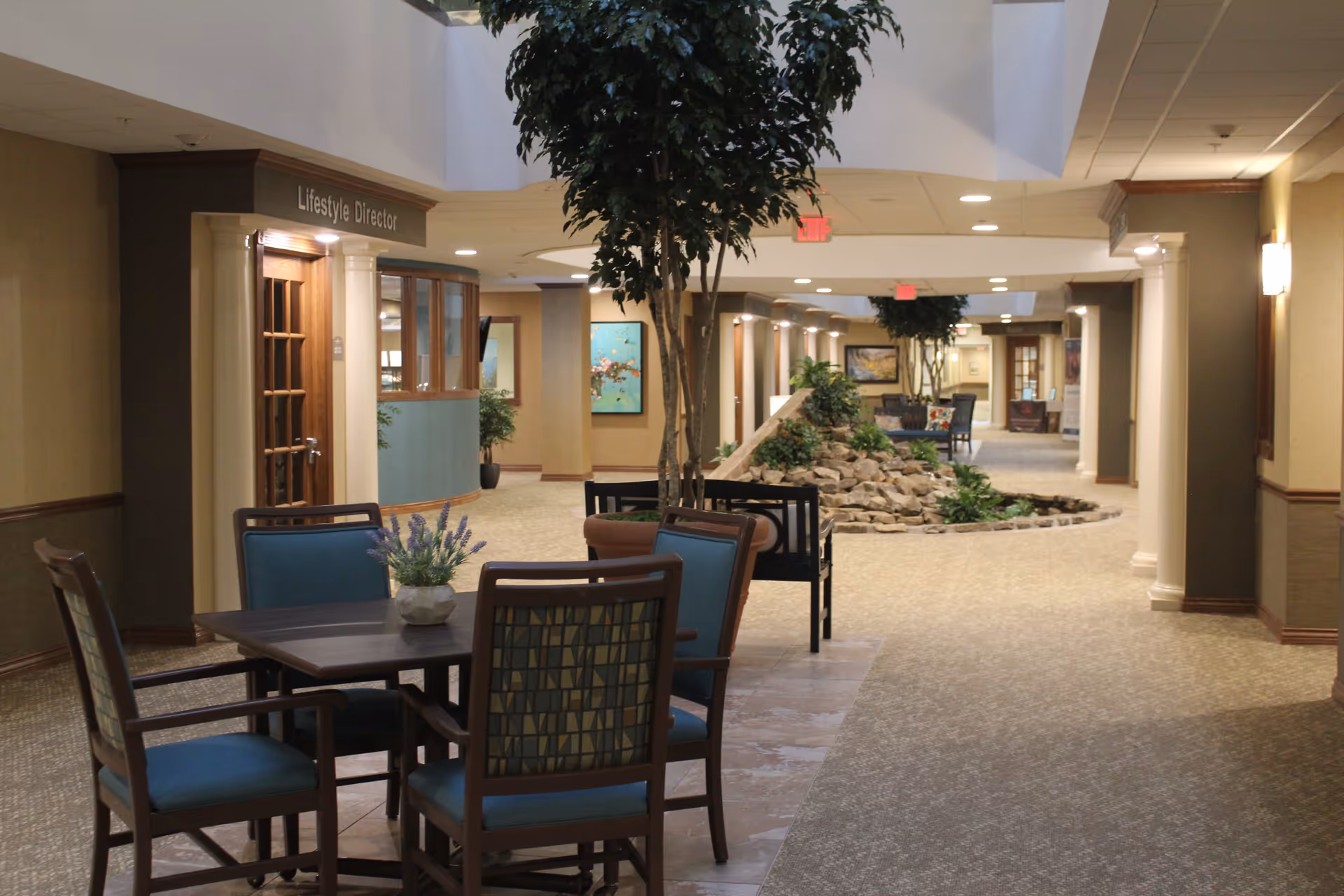 Interior view of a retirement community hallway with a seating area featuring a table and four chairs with blue cushions. The hallway has beige walls, carpeted floors, potted plants, and a rock garden centerpiece. A door labeled 'Lifestyle Director' is visible on the left side.