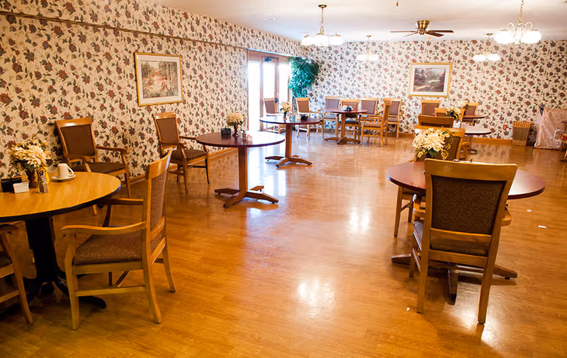 A dining room with wooden tables and chairs arranged neatly on a polished wooden floor. The walls are covered with floral wallpaper and decorated with framed paintings. There are flower arrangements on the tables and ceiling lights providing warm illumination. Large windows with curtains allow natural light into the room.