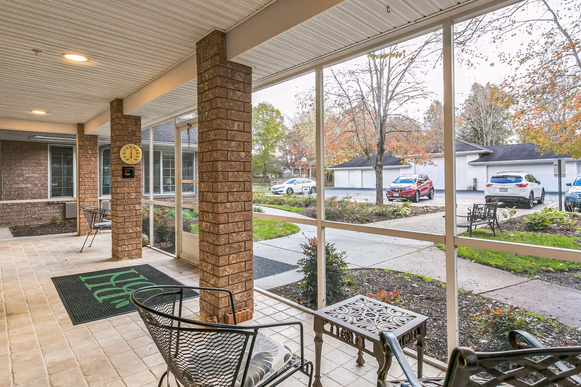 A covered outdoor patio area with brick pillars and a tiled floor, furnished with metal chairs and a small decorative table. The patio is enclosed with large screened windows, overlooking a parking area with several cars and trees with autumn foliage.