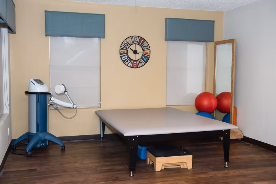 A therapy or exercise room with a large padded table, a small wooden step stool underneath, two windows with blue valances, a colorful wall clock, a full-length mirror, and several exercise balls in red and blue colors. There is also a piece of medical or therapy equipment on wheels positioned near one window.