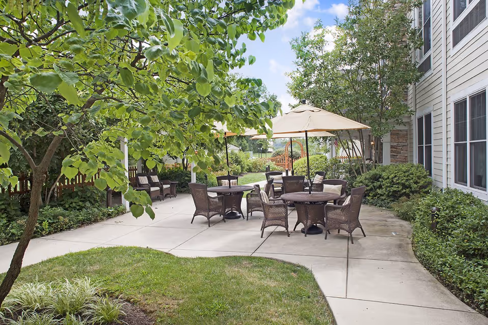 Outdoor patio area at Sunrise of Dresher with several round tables and wicker chairs, some shaded by large beige umbrellas. The patio is surrounded by green trees, bushes, and a well-maintained lawn, with the building visible on the right side under a partly cloudy sky.