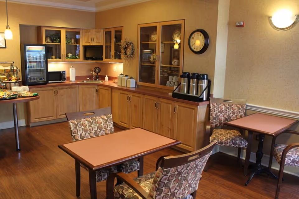 A cozy dining area in an assisted living facility featuring wooden tables and cushioned chairs with patterned upholstery. The room includes a kitchenette with wooden cabinets, a small refrigerator, a microwave, coffee dispensers, and a clock on the wall. The space is warmly lit with pendant and wall lights.