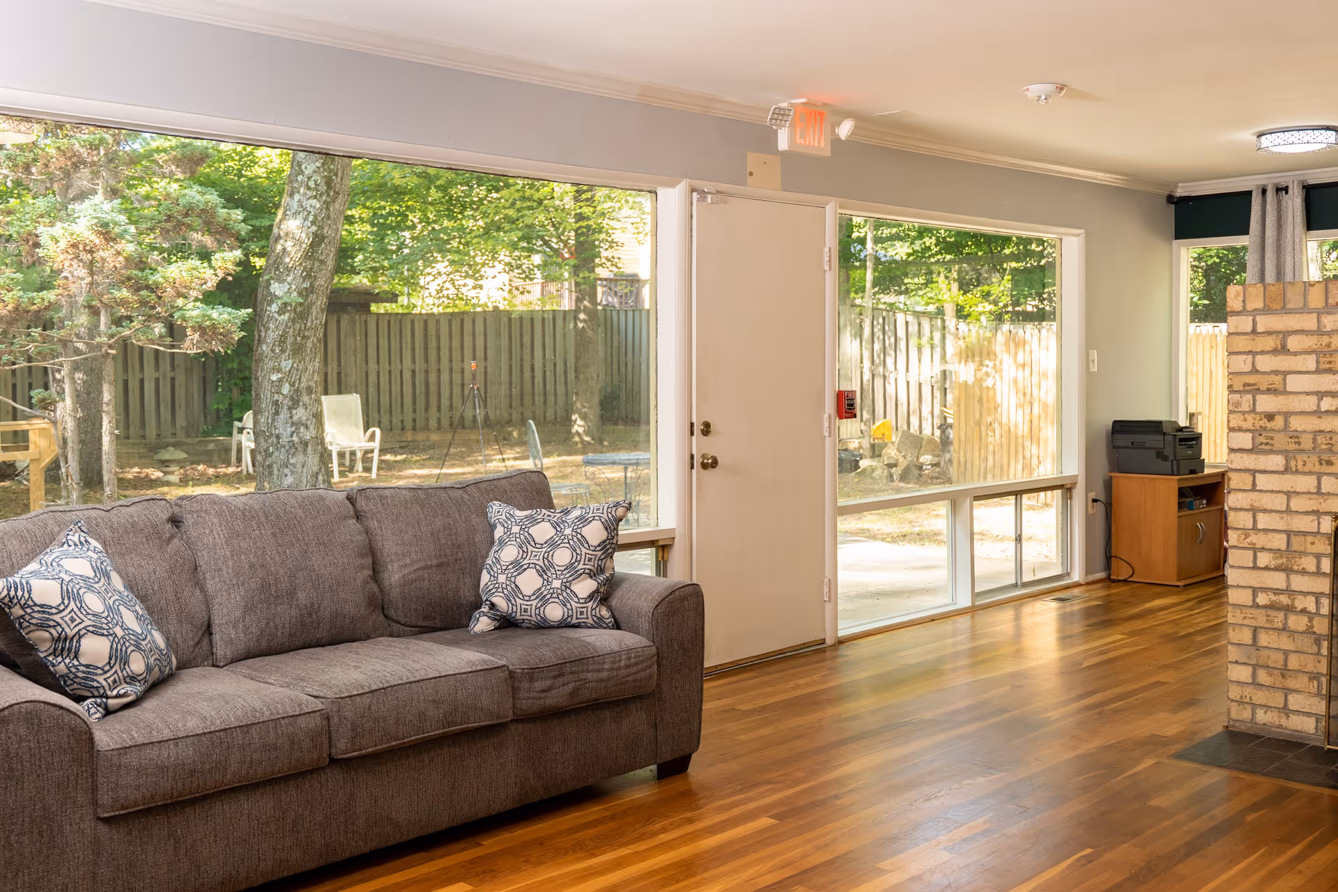 A living room with a gray couch adorned with two patterned pillows, large windows showing a fenced backyard with trees and outdoor chairs, a wooden floor, and a brick fireplace partially visible on the right.