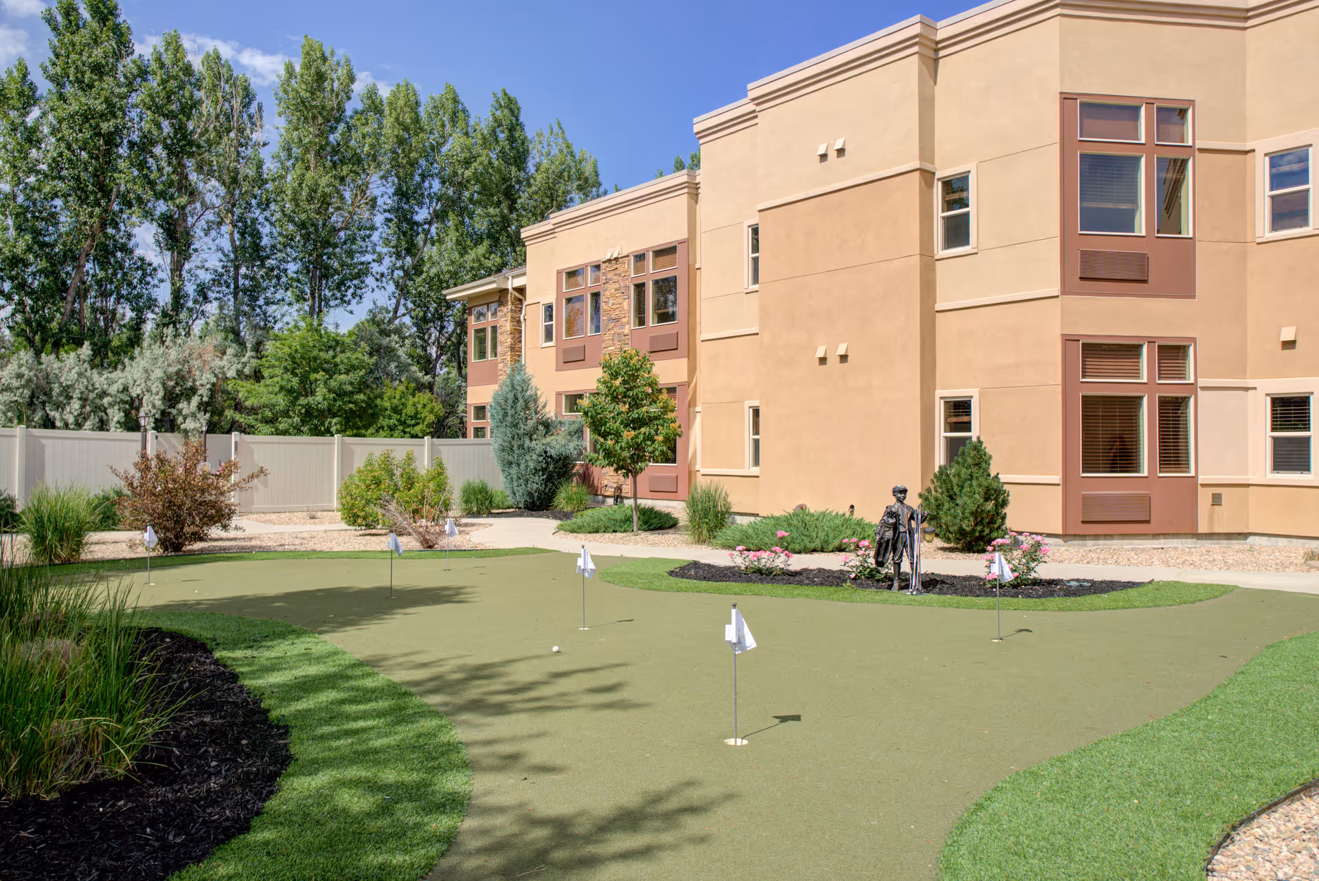 Landscaped courtyard with a small putting green in front of a beige two-story senior living building under a clear blue sky.