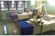 Two neatly made hospital beds with medical equipment and bedside tables in a pale green patient room.
