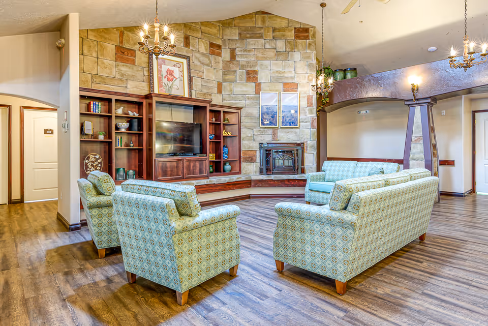 A spacious living room with patterned light blue armchairs and sofas arranged around a wooden entertainment center with a TV. The back wall features a stone facade with a fireplace and framed artwork above it. The room has wooden flooring, warm lighting from chandeliers, and an archway leading to other areas.