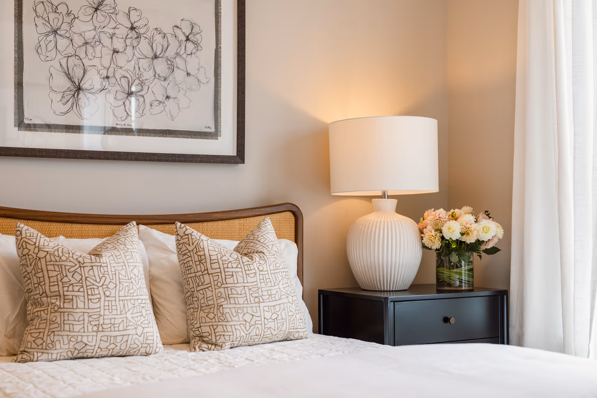 A cozy bedroom corner featuring a bed with two patterned pillows and white bedding. Next to the bed is a black nightstand with a white textured lamp and a glass vase filled with light pink and white flowers. Above the bed hangs a framed artwork of abstract black floral sketches on a white background. Light beige walls and white curtains complete the serene setting.