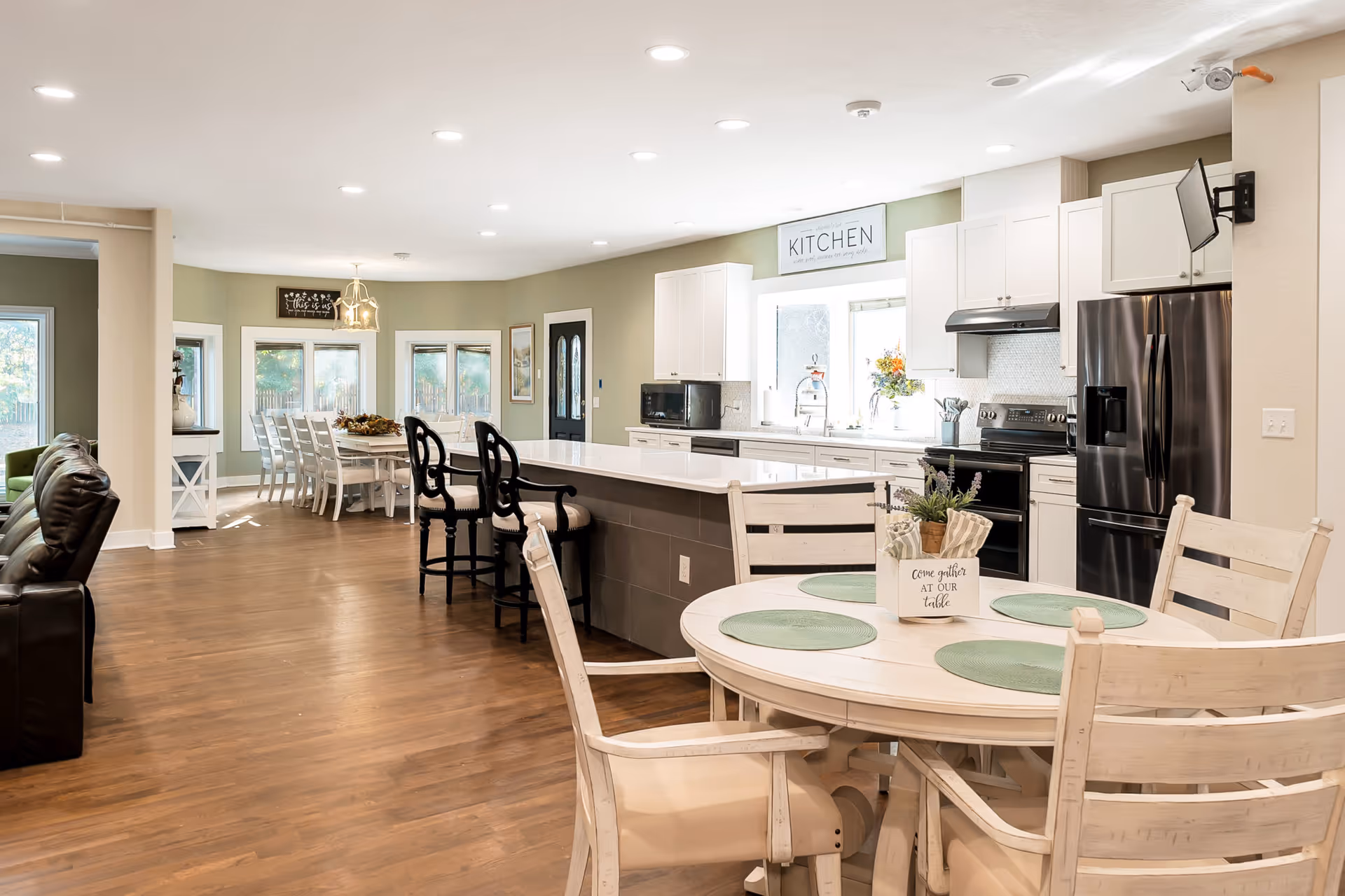 A bright, open kitchen and dining area in a senior living facility. The kitchen features white cabinets, a black refrigerator, stove, microwave, and a large island with bar stools. In the foreground, there is a round dining table with four chairs and green placemats. In the background, there is a larger dining table with white chairs near windows, and a living area with brown leather chairs to the left. The walls are painted light green and beige, and the floor is wood.