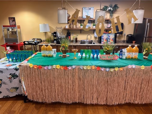 A decorated indoor tiki bar setup with a grass skirt table covering and colorful flower garland. The table is covered with a green cloth and holds several green and blue glasses, bottles of orange juice, and small decorative plants. Behind the table is a kitchen counter with a sink, coffee maker, and various kitchen items. Above the counter hangs a banner spelling 'ALOHA' in gold letters.