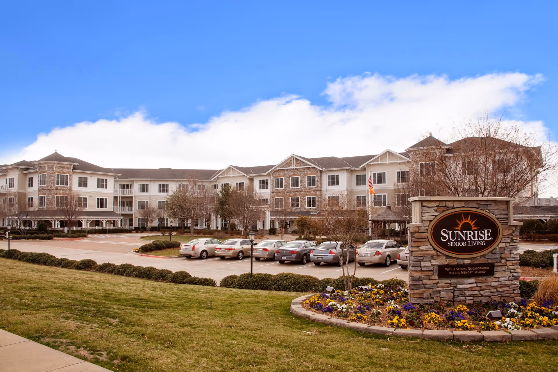 Exterior view of Sunrise Senior Living facility in Plano, showing a large three-story building with stone and white siding, a parking lot with several cars, a landscaped area with grass and flowers, and a stone sign with the Sunrise Senior Living logo.