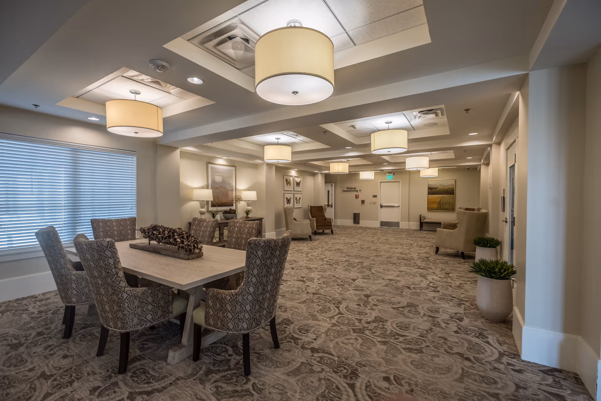 A spacious, well-lit common area in a senior living facility featuring a large patterned carpet, a wooden table with six upholstered chairs, several armchairs along the walls, decorative plants in pots, framed artwork, and multiple ceiling lights.