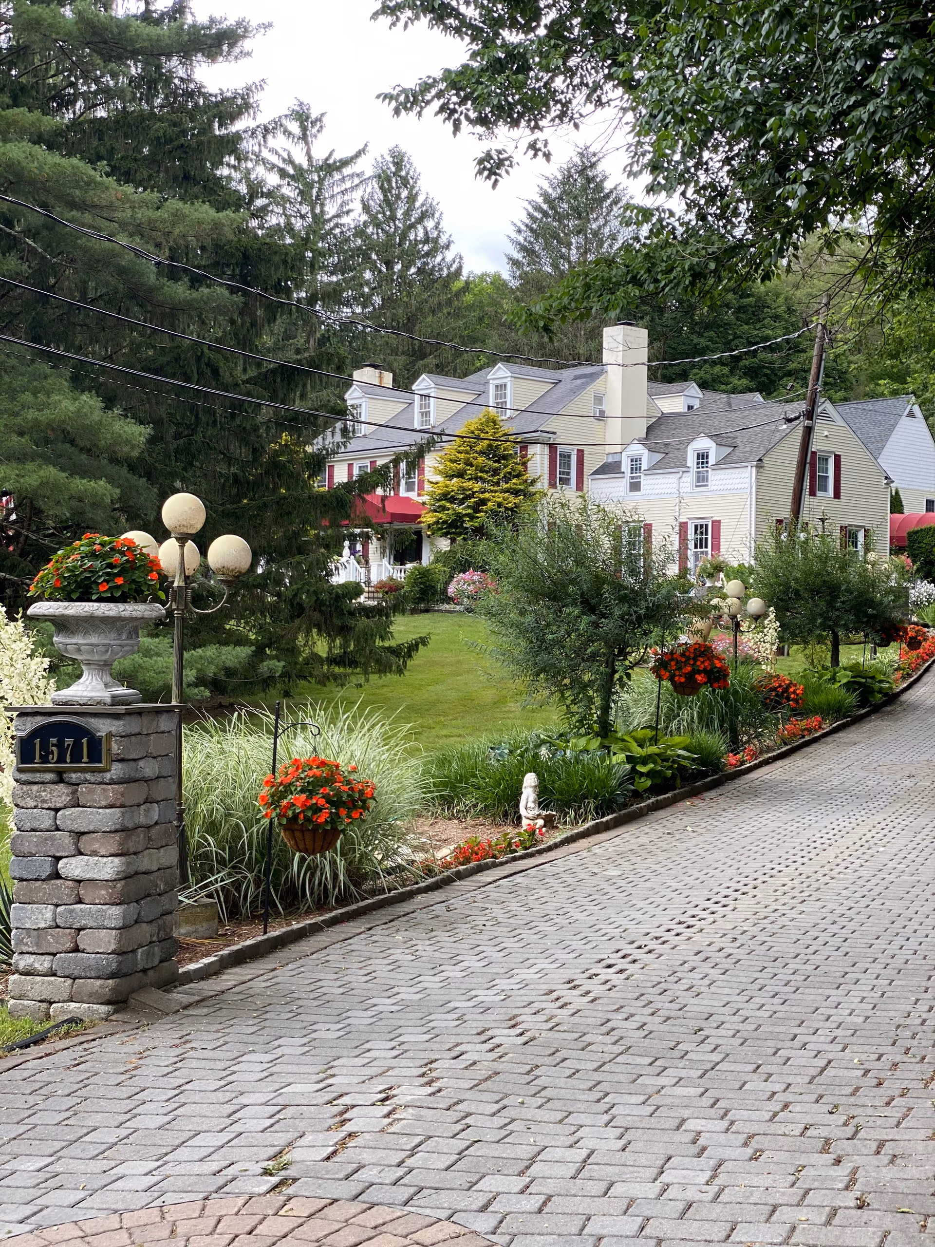 A paved driveway leading to a large, multi-story colonial-style building with cream-colored siding and red shutters, surrounded by lush greenery, trees, and colorful flower beds. There is a stone pillar with the number 1571 and decorative flower pots near the entrance.