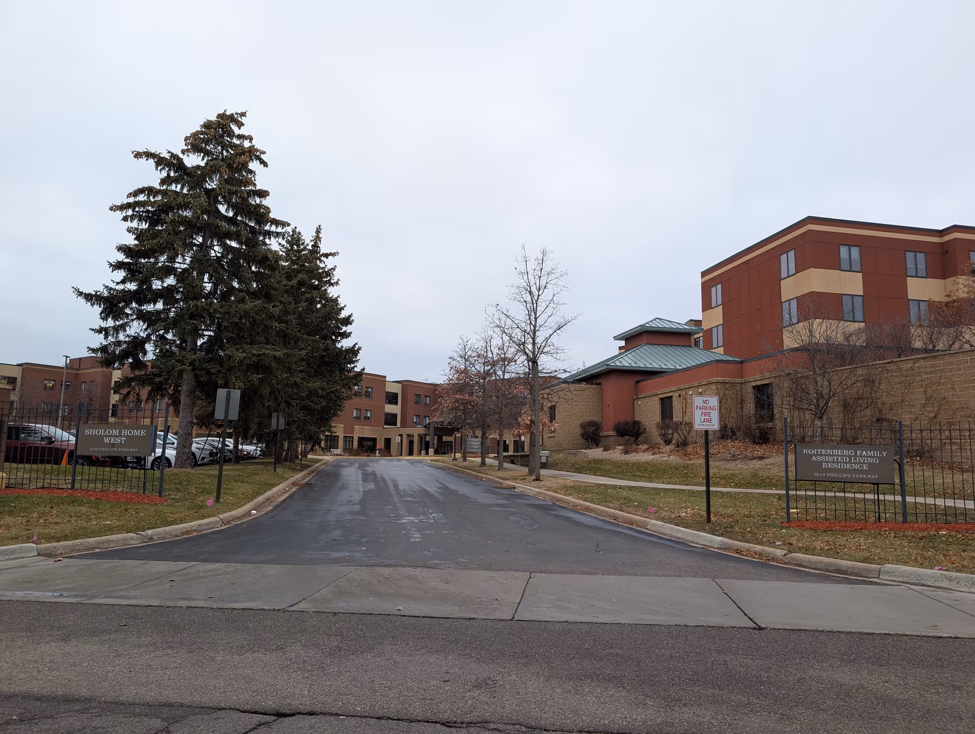 Driveway leading to the front entrance of a multi-story assisted living building with signs, trees, and parked cars.