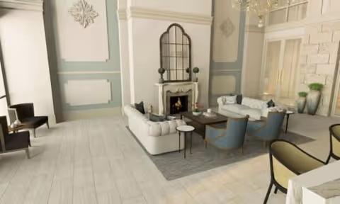 A spacious and elegant living room with light-colored wooden flooring, a white fireplace with a mirror above it, two white sofas, two blue armchairs, a dark wooden coffee table, and decorative plants. The room features high ceilings, large windows, and soft pastel wall colors with decorative molding.
