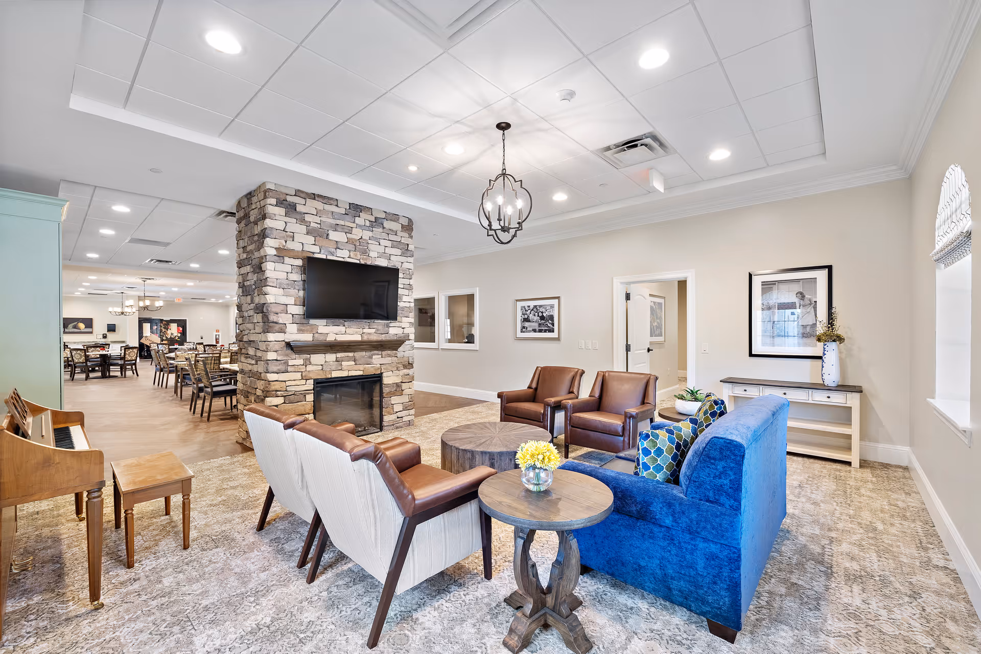 A bright and spacious living room area in Worthington Manor featuring a stone fireplace with a mounted flat-screen TV, a blue sofa with patterned pillows, four armchairs arranged around two round wooden tables, a small wooden piano with a stool, and a dining area visible in the background.