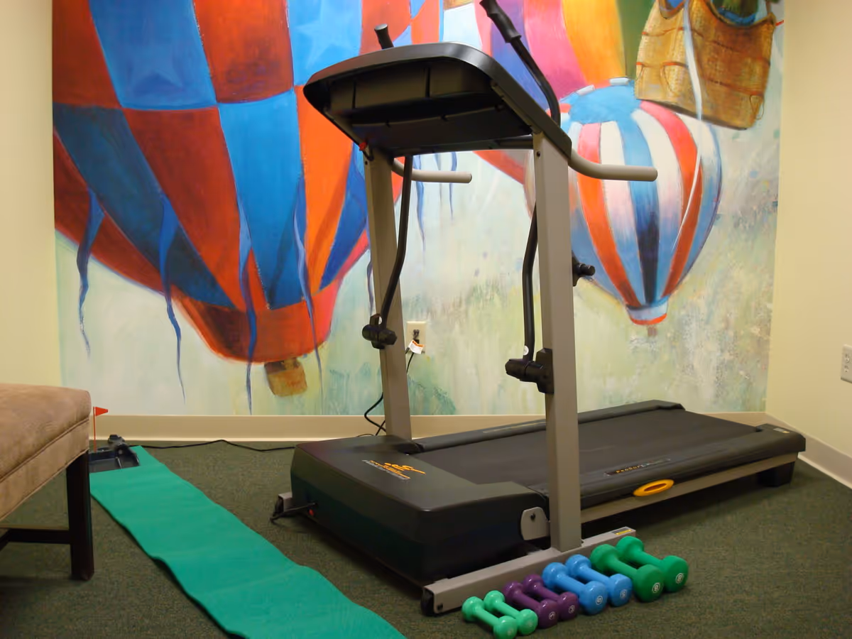 Small exercise room with a treadmill, colorful hot-air-balloon mural on the wall, a green exercise mat and several dumbbells.