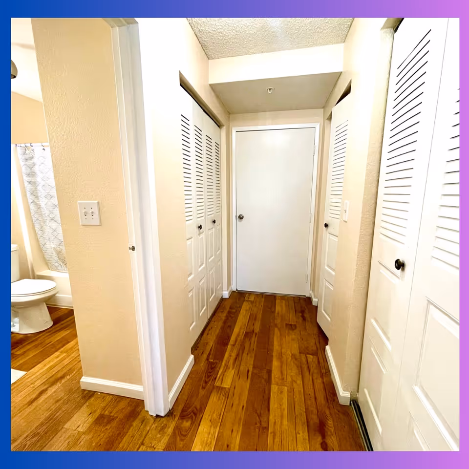 Narrow interior hallway with hardwood floors, multiple white louvered closet doors, a closed door at the end, and a bathroom visible on the left.