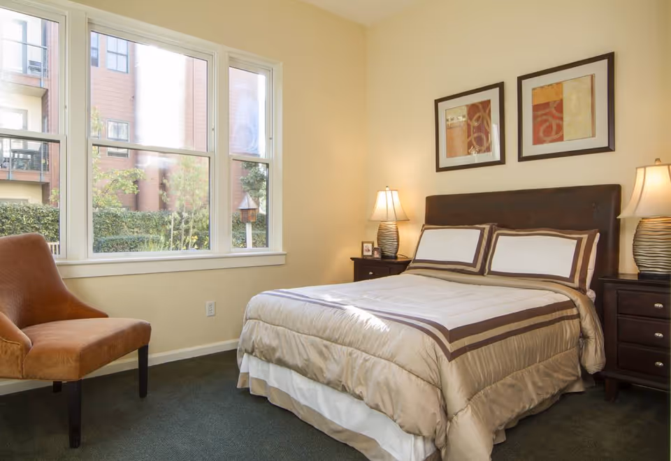 A bedroom with a neatly made bed featuring beige and brown bedding. There are two matching bedside tables with lamps on each side of the bed. A brown upholstered chair is positioned near a large window that lets in natural light. The walls are painted a light beige color and two framed abstract artworks hang above the bed.
