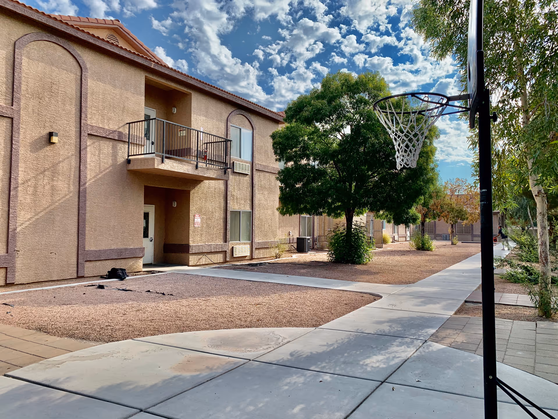 Outdoor view of Sana Assisted Living facility showing a two-story beige building with a balcony, several windows, and a door. There is a basketball hoop on the right side near a paved walkway, and a large green tree in the center of the courtyard area with gravel ground cover. The sky is partly cloudy.