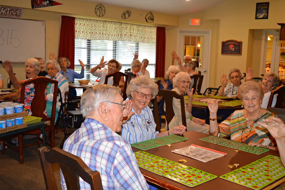 A group of senior residents sit at tables in a communal room smiling and raising their hands while playing bingo.