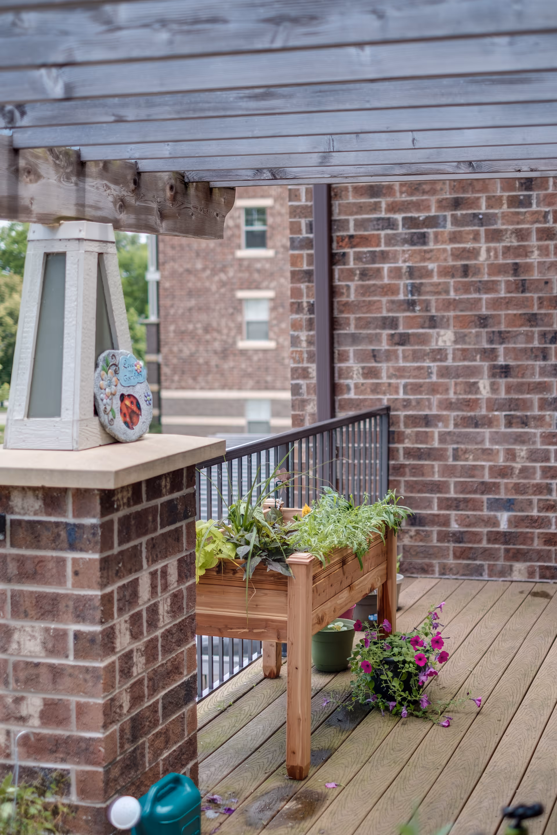 A wooden balcony or patio area with a raised wooden planter box filled with green plants and a pot of purple flowers on the wooden floor. The balcony has a brick pillar and railing, with a wooden pergola overhead. A decorative stone with a ladybug design is placed on the pillar.