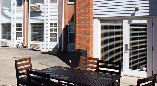 Outdoor patio area at Lincoln Village with a metal table and several chairs on a concrete surface, adjacent to a building with white siding and brick walls, featuring windows and a glass door with blinds.
