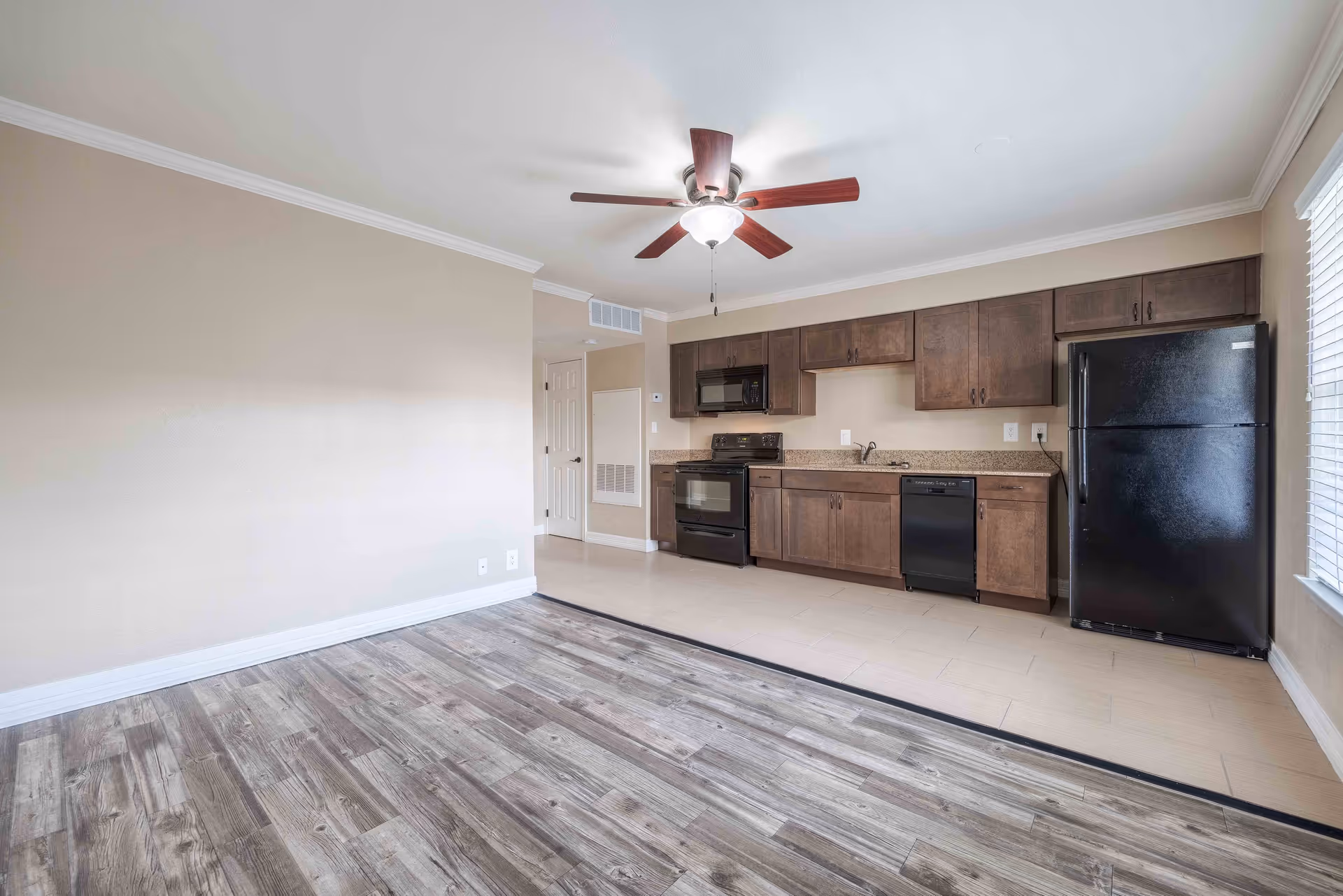 An empty kitchen area in a senior living facility with dark wood cabinets, black appliances including a refrigerator, stove, microwave, and dishwasher. The kitchen has a beige countertop and beige tiled floor, transitioning to a wood-patterned floor in the adjacent living space. A ceiling fan with wooden blades and a light fixture is mounted on the white ceiling. There is a window with white blinds on the right side.