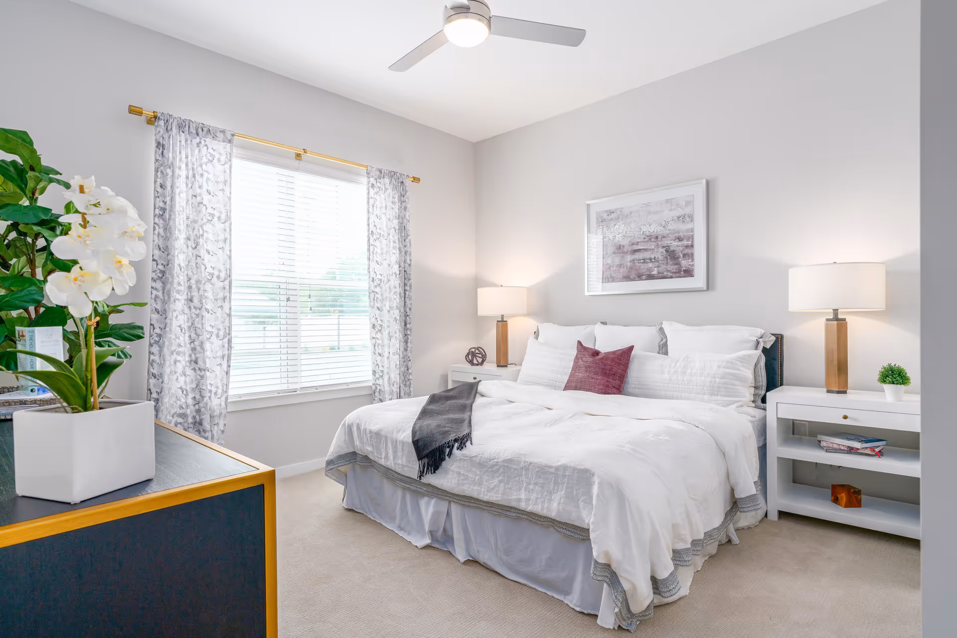 A bright and neatly arranged bedroom with a large bed featuring white bedding and a purple accent pillow. There are two white nightstands on either side of the bed, each with a lamp and decorative items. A window with patterned curtains allows natural light to fill the room. A ceiling fan with a light is mounted on the ceiling, and a potted plant sits on a dark dresser with gold trim.