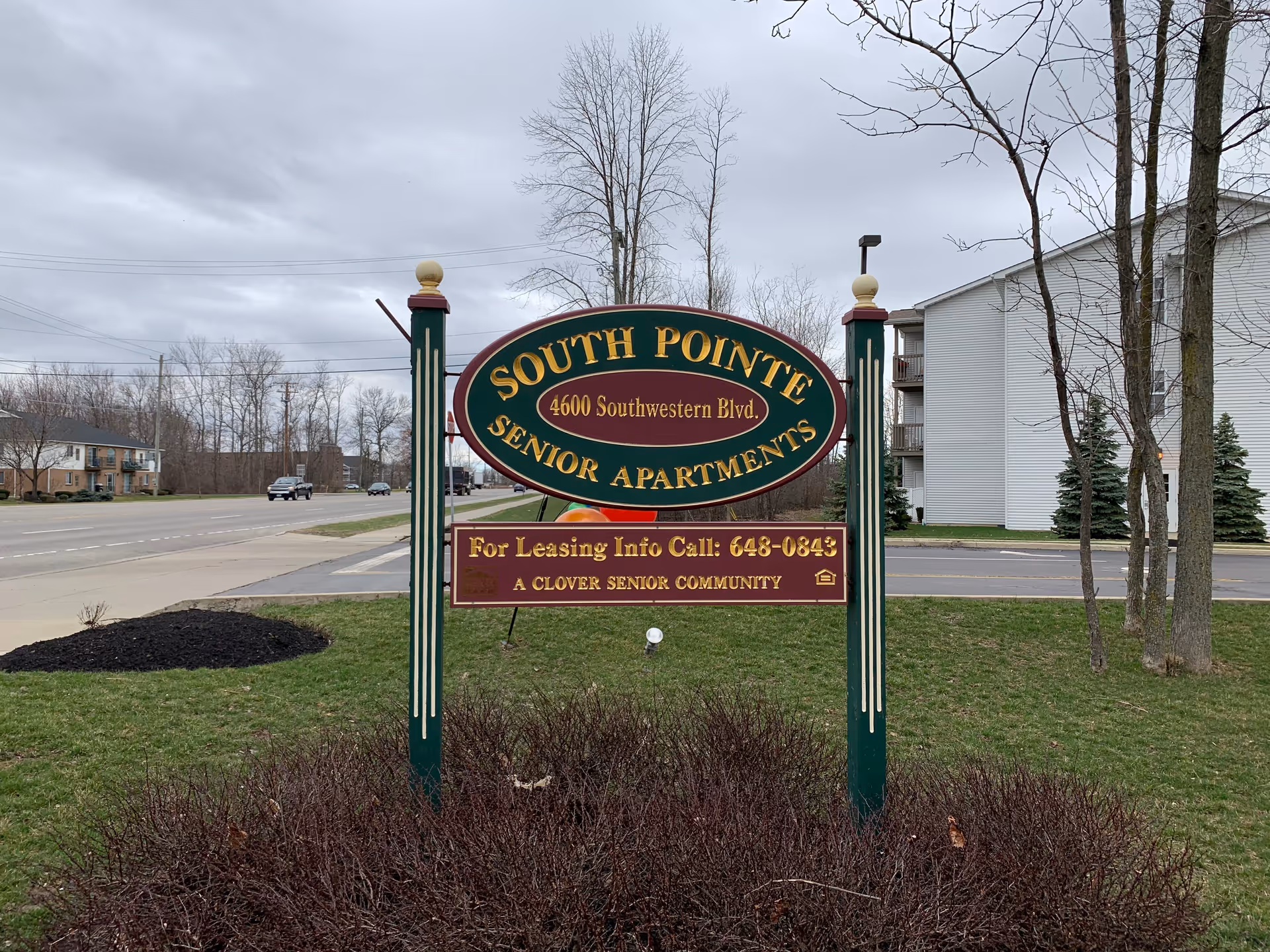 A roadside sign reading 'South Pointe Senior Apartments' on a grassy lawn with the apartment building and a street in the background.