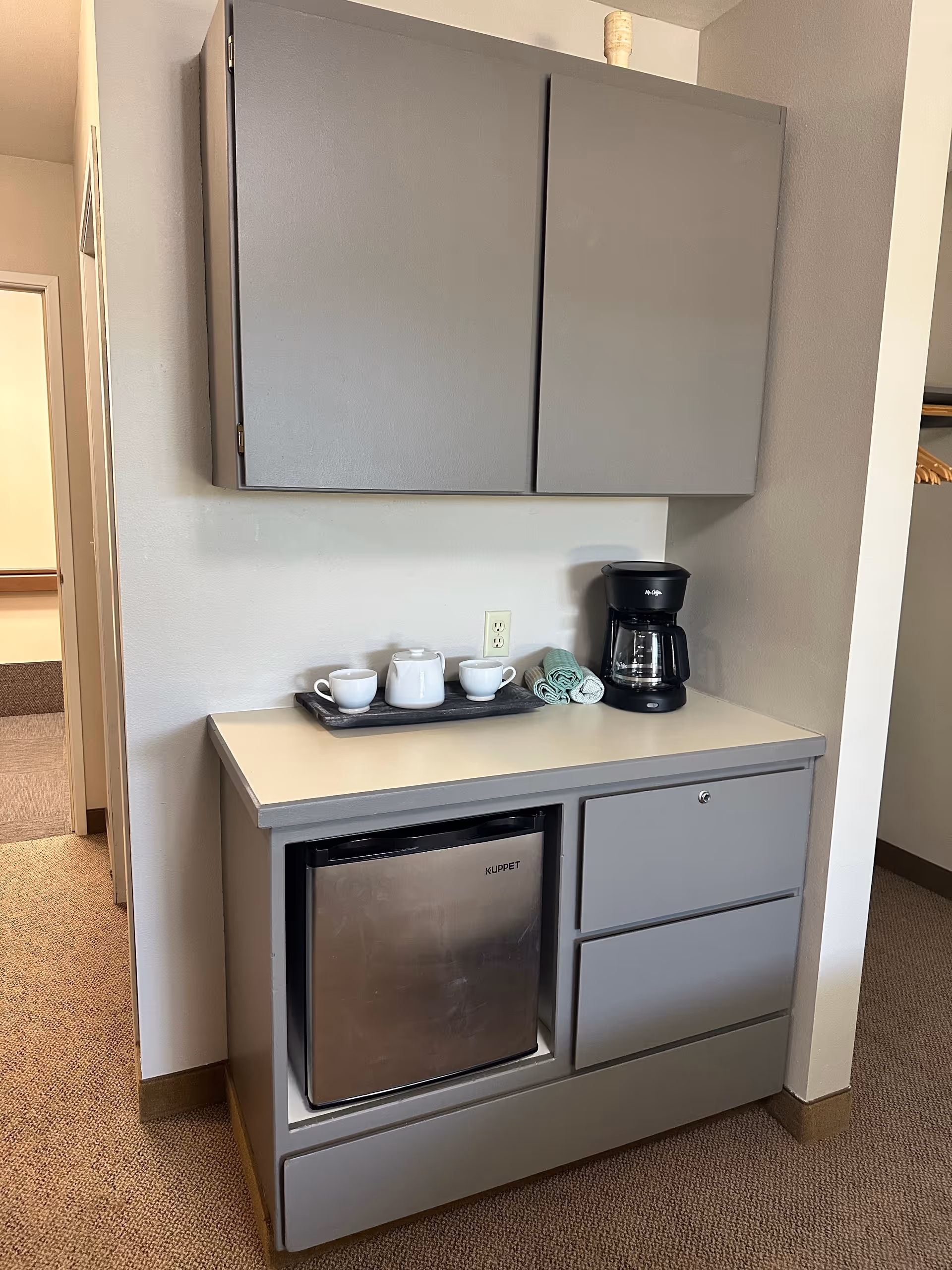 Small kitchenette with gray cabinets, a mini fridge, coffee maker and a tea set on the countertop.