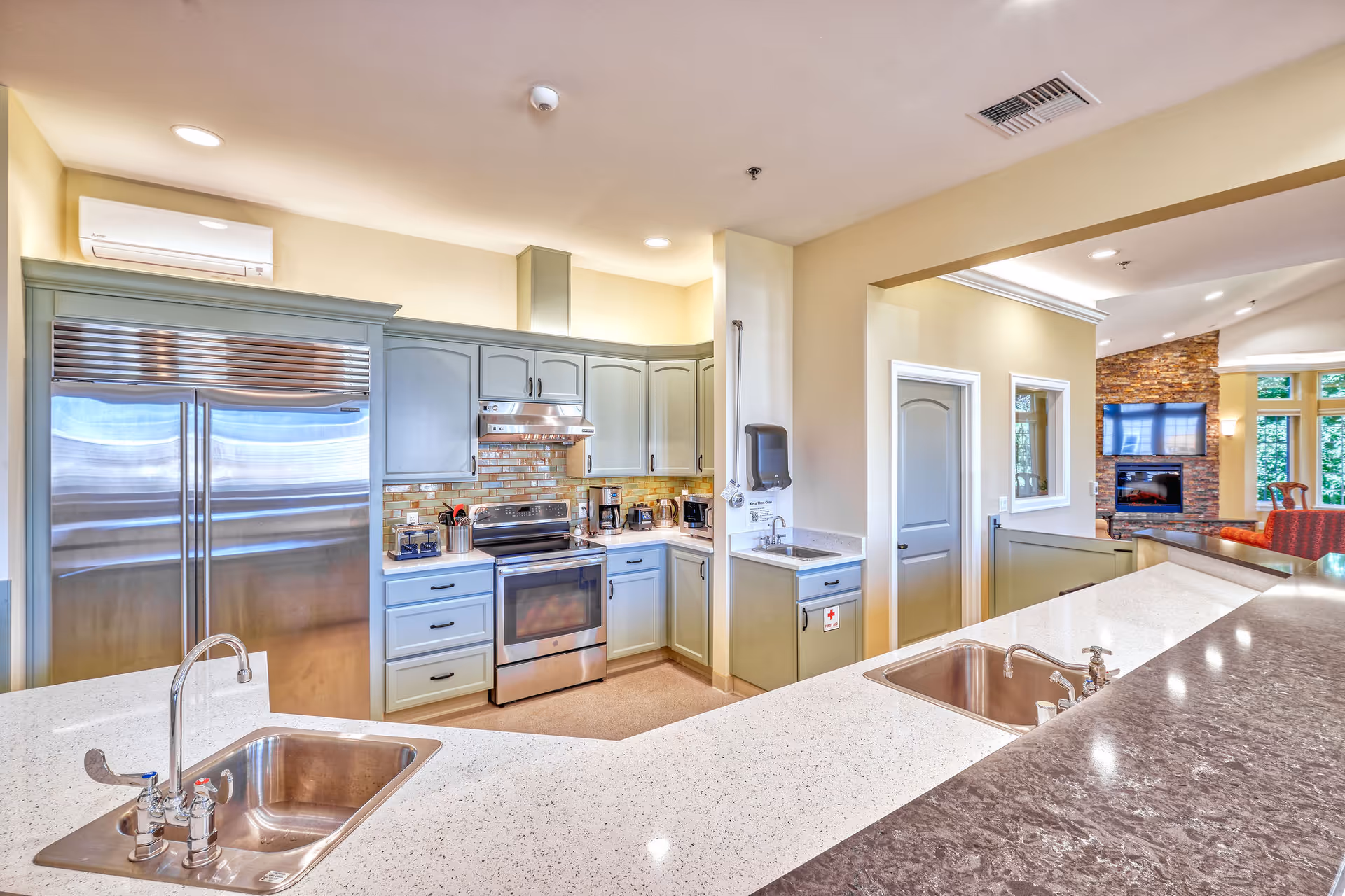 A modern kitchen area with light green cabinets, stainless steel refrigerator and oven, a brick backsplash, and two sinks set in white countertops. The kitchen opens into a living area with a stone fireplace and large windows letting in natural light.