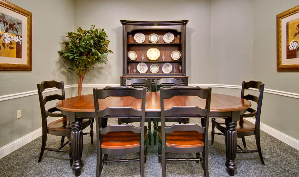 A formal dining room with a wooden oval table, six chairs, a china cabinet displaying plates, and framed wall art.