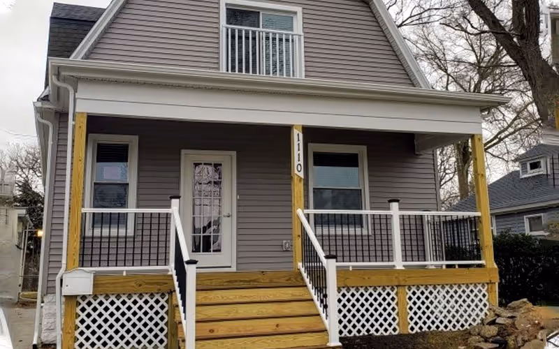 Two-story residential building with a covered front porch, railing, and wooden stairs.