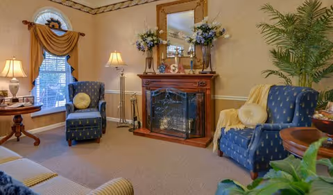 A cozy living room with two blue armchairs, a wooden fireplace with a decorative screen, a large mirror above the fireplace, floral arrangements, a window with a brown valance, a floor lamp, and a round wooden side table with a lamp and decorative items.