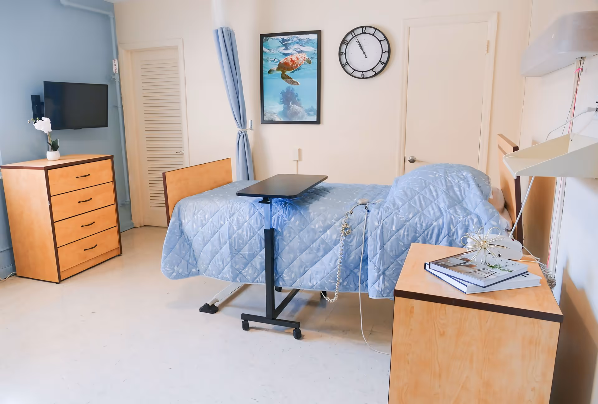 A senior care facility bedroom with a hospital-style bed covered in a light blue quilted blanket. There is an overbed table positioned over the bed, a wooden dresser with a white orchid plant on top, a wall-mounted TV, a clock showing 12:05, a framed picture of a sea turtle swimming underwater, and a wooden nightstand with books and a decorative item. The room has light-colored walls and floor, and a blue privacy curtain is partially visible.