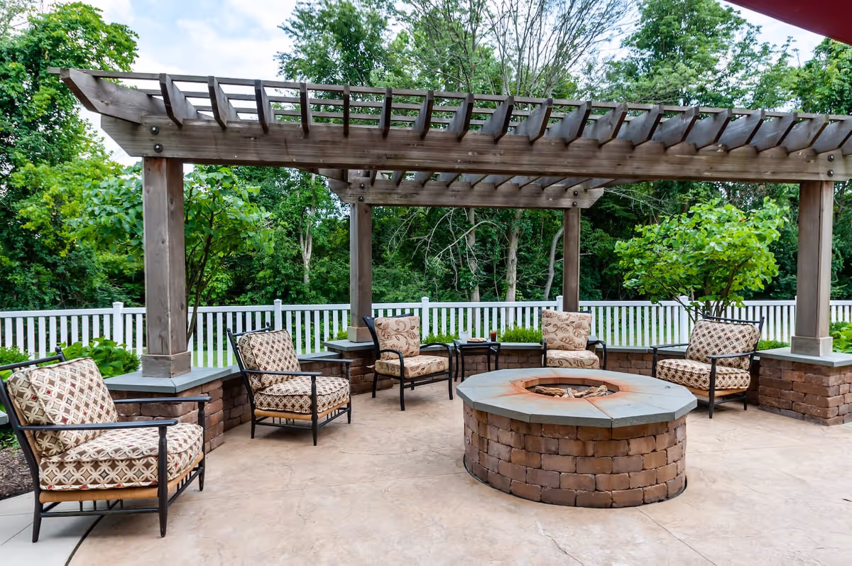 Outdoor seating area with five cushioned chairs arranged around a circular brick fire pit under a wooden pergola, surrounded by greenery and a white picket fence.