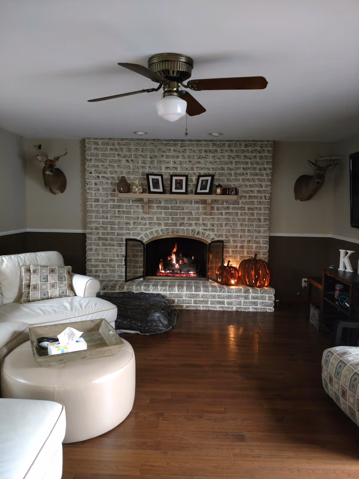 Cozy living room with a white sectional sofa, a round ottoman with a wooden tray holding a tissue box, a brick fireplace with a fire burning inside, and two mounted deer heads on the walls. There is a ceiling fan with wooden blades, framed photos and decorative items on the fireplace mantel, and pumpkin decorations near the fireplace.