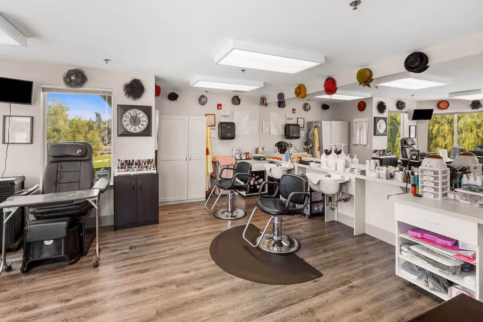 Interior view of a senior living community salon with multiple black salon chairs, hair washing stations, nail polish display, and various hair care products on counters. The room has large windows showing greenery outside, hats hanging on the walls, and a clock on the wall.