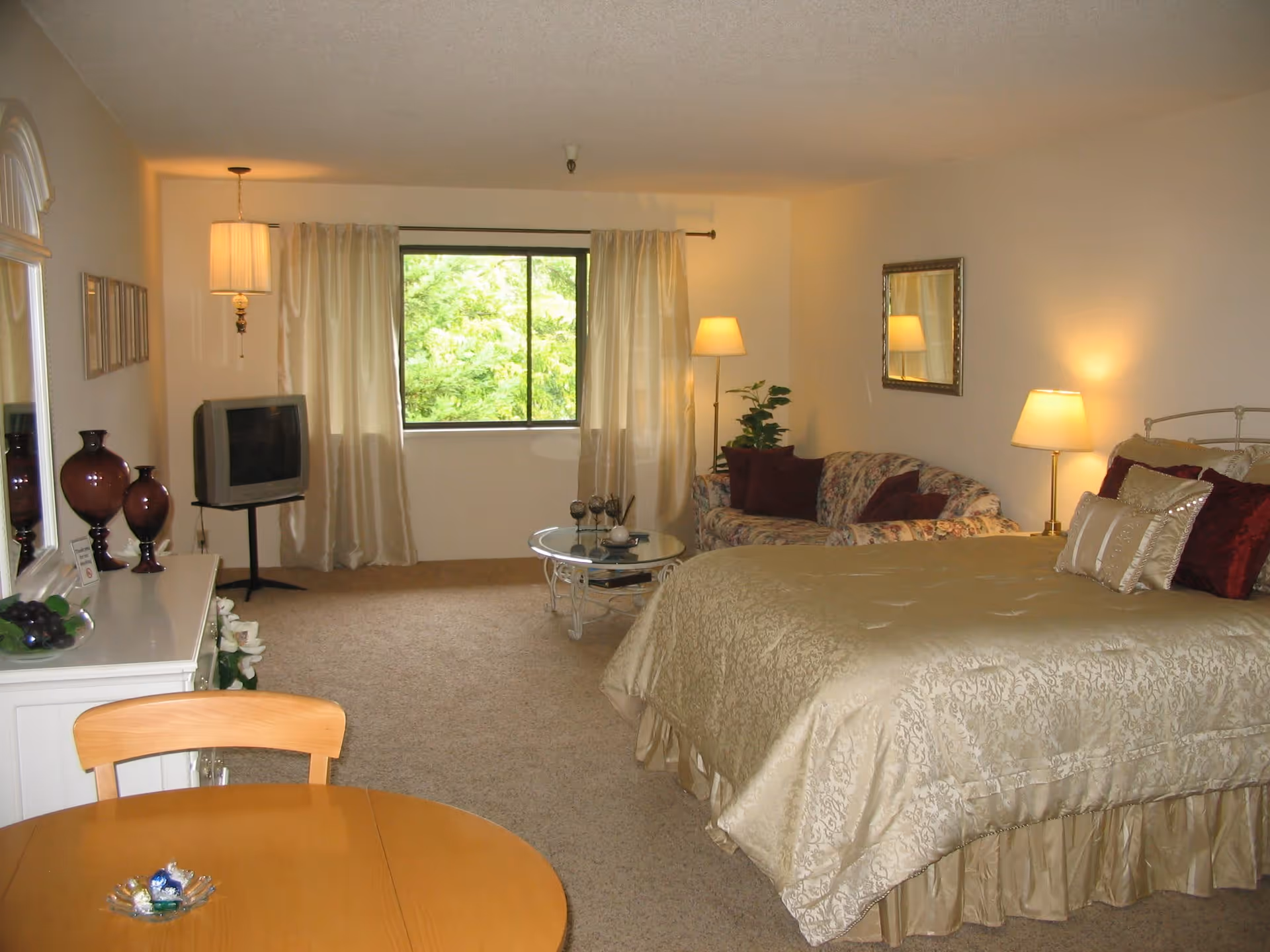 A cozy retirement community room featuring a large bed with gold bedding and multiple pillows, a floral-patterned sofa with burgundy cushions, a round glass coffee table, a wooden dining table with chairs, a TV on a stand, and a window with cream-colored curtains letting in natural light. The room is softly lit by several lamps and decorated with vases and a mirror on the wall.